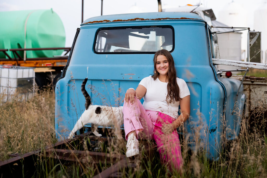 A young woman wearing a white t and pink pants is seated on the rusted frame of a blue pickup truck on her family's property for her North Dakota senior photos.