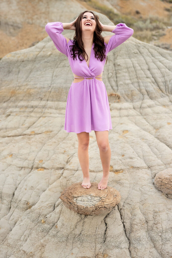 An image from a Williston senior photo gallery depicts a senior girl wearing a purple dress tousles her hair as she stands barefoot atop a butte at the American Legion Badlands.