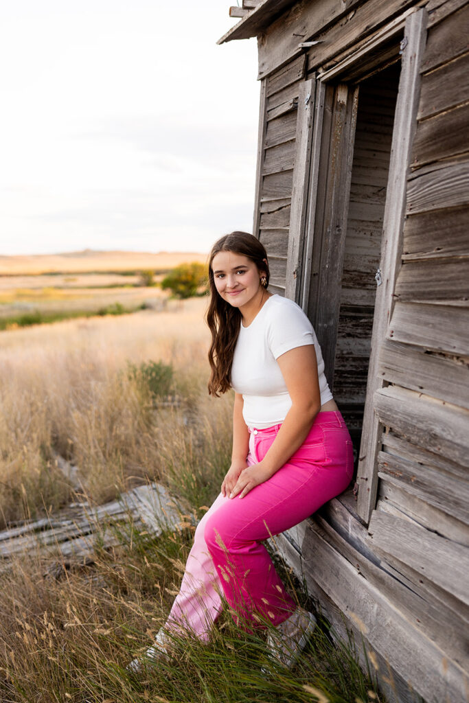 A young woman wearing a white t and pink pants is seated on the window sill of a shed on her family's property for her North Dakota senior photos.