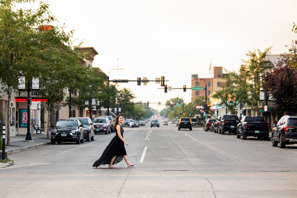 An image from a Williston senior session with North Dakota senior photographer, Kellie Rochelle Photography, is pictured.  A young woman with dark hair walks barefoot across the street in downtown Williston in a black dress.