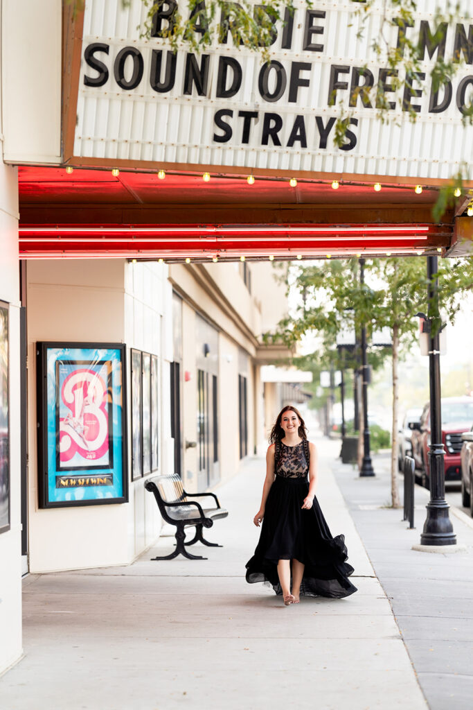 A North Dakota high school senior girl wearing a long, black dress walks under the marquee at the movie theater during a Downtown Williston senior photo session with Kellie Rochelle Photography.