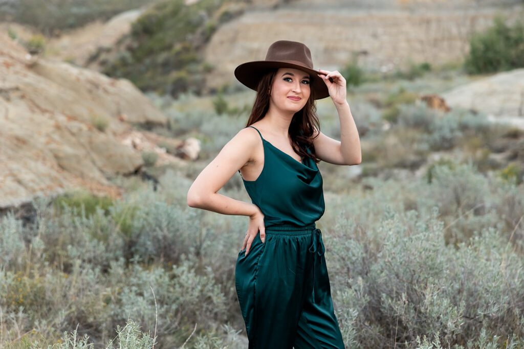 An image from a Williston senior photo gallery depicts a senior girl wearing a deep green ensemble touches the brim of her brown hat stands in a field at the American Legion Badlands.