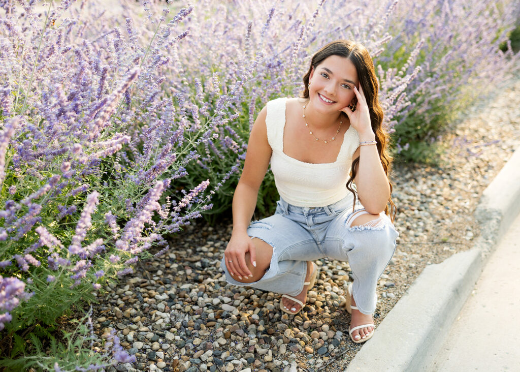 Crouching in ripped jeans and a cream body suit near purple flowers, a Williston senior girl poses for her senior photos with North Dakota photographer Kellie Rochelle Photography.