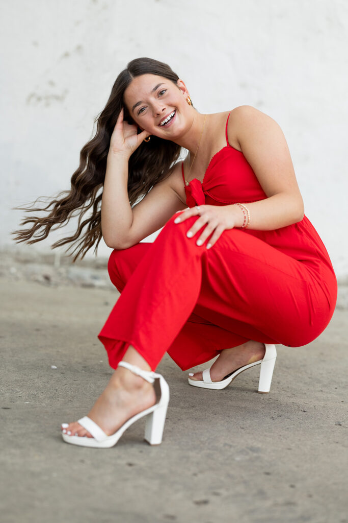 A young woman with long, dark hair wearing a red romper and white heels smiles and crouches during a downtown Williston senior photo session with Kelle Rochelle Photography