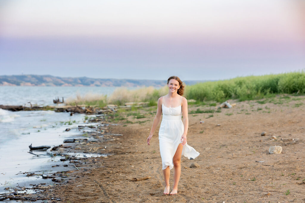 A young woman wearing a white dress blowing in the breeze walks barefoot along the shoreline at Lewis and Clark State Park with a lavender sunset peeks over the horizon in the background during a shoot with North Dakota senior photographer, Kellie Rochelle Photography.