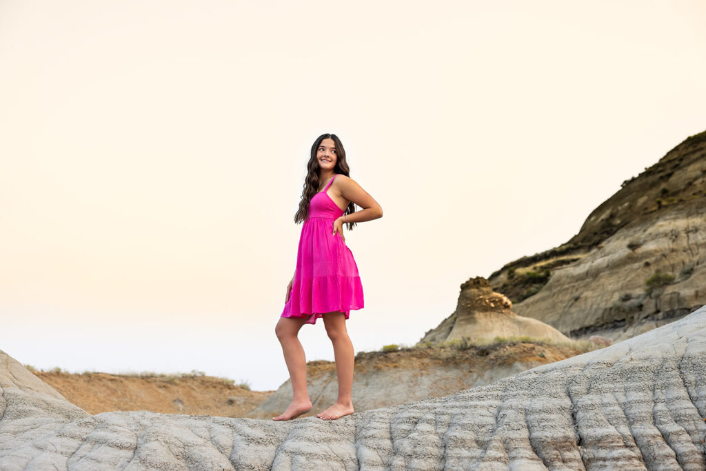 An image from a Williston senior photo gallery depicts a senior girl wearing a hot pink dress stands barefoot atop a butte at the American Legion Badlands.