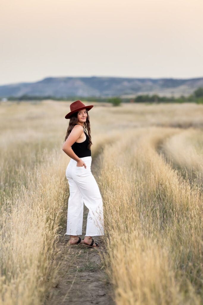 A senior picture taken at the American Legion Badlands outside Williston, ND is shown depicting a young woman in white raw hemmed pants and a black body suit wearing a rust colored hat stands with her hands in her back pockets on a path at the American Legion Badlands. 