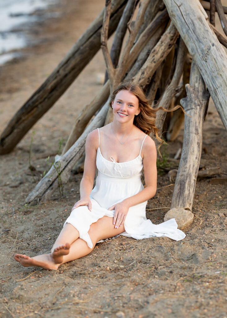 A Williston ND high school senior is pictured wearing a flowy white dress as she is seated against a stack of timber near the shoreline at Lewis and Clark State Park 