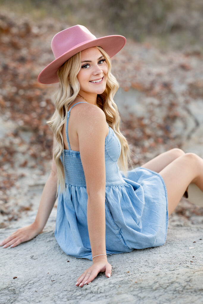 Wearing a denim dress and pink hat, a blonde high school senior girl is photographed seated on the rocky ground at Lewis & Clark State Park during a senior session with Kellie Rochelle Photography.