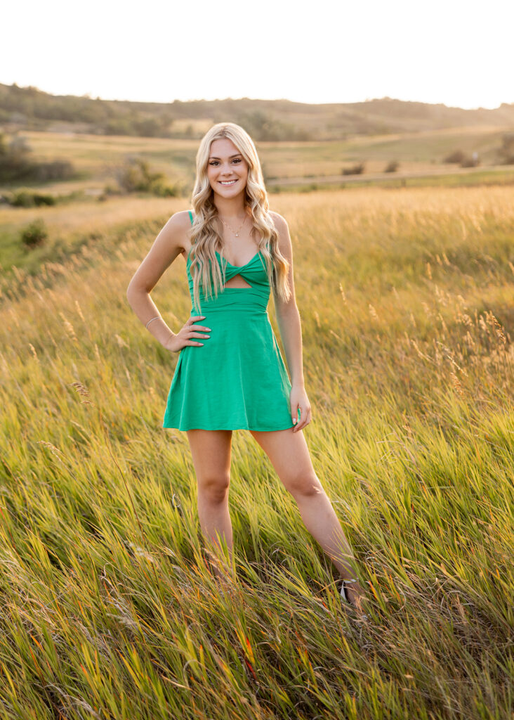 An image from a Williston senior photo session is shown. A blonde young woman wearing a green dress is pictured in a grassy field at golden hour at Lewis & Clark State Park.