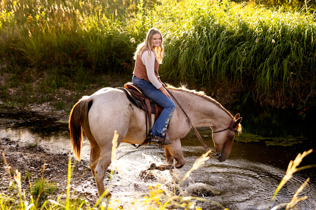 A blonde young woman is seated on the saddle of her brown horse who walks through a creek on her family's North Dakota ranch for her North Dakota senior pictures.