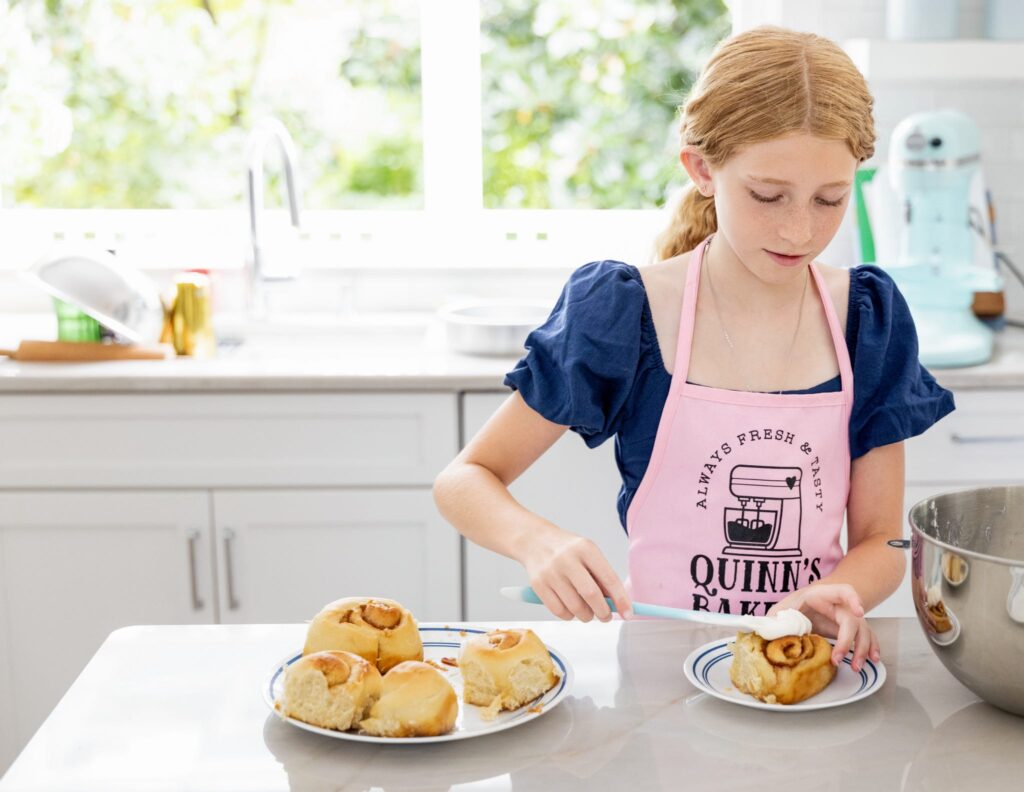 A young girl wearing a pink apron that reads,' Quinn's Bakery' frosts a cinnamon roll in the brightly lit kitchen of her Washington D.C. home during a family videography session with Kellie Llewellyn, a North Dakota recipient of Backing Small Businesses grant.