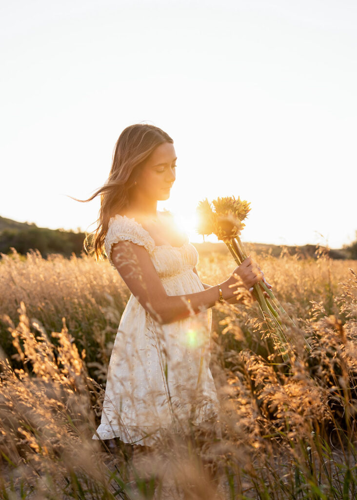 graduate standing in a field of sunflowers at golden hour with the glow of the sun behind her