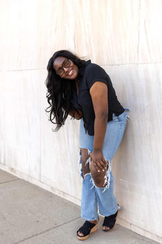 Destiny leaning against light stone column in black blouse and jeans