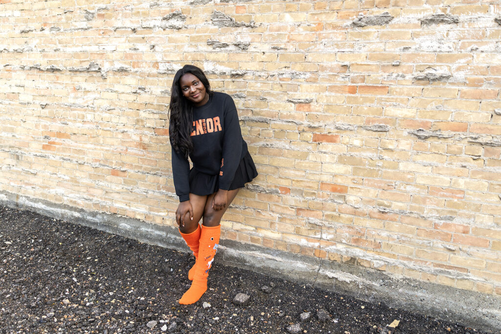 Williston senior girl in “Senior 2025” sweatshirt and orange socks sitting by brick wall