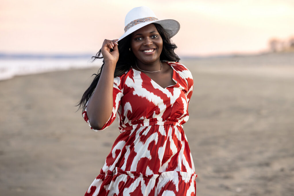 Senior girl in red and white dress with cowboy hat leaning on driftwood at lake