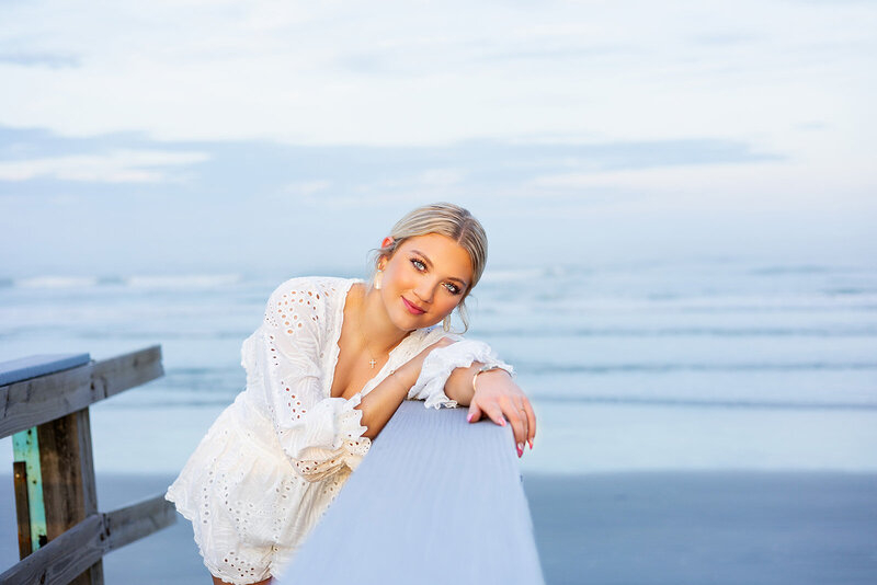 A soft, golden glow frames a young woman leaning gently on a boardwalk railing by the sea.








