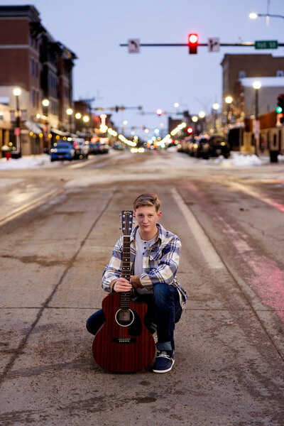 Teen with guitar kneels in glowing street, city lights sparkle.