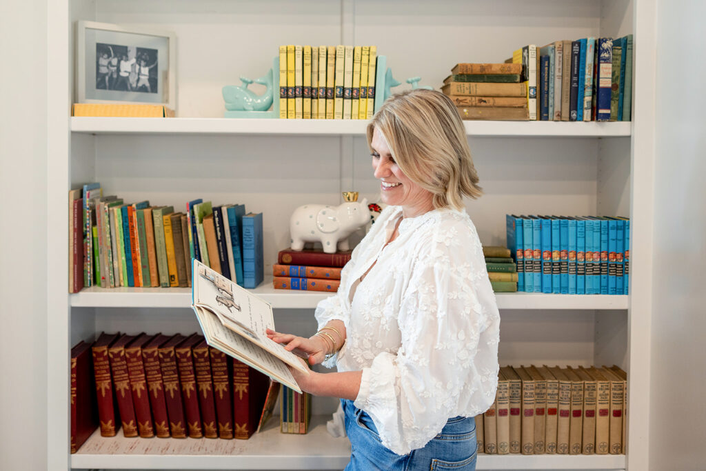 A blonde woman wearing a white blouse and jeans flips through the pages of a book before a bookshelf during an Austin TX branding session