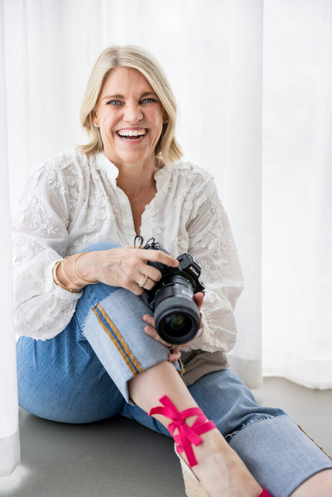 A blonde photographer wearing hot pink wedges holds her Nikon camera and smiles for a headshot before a white backdrop during an Austin TX branding session