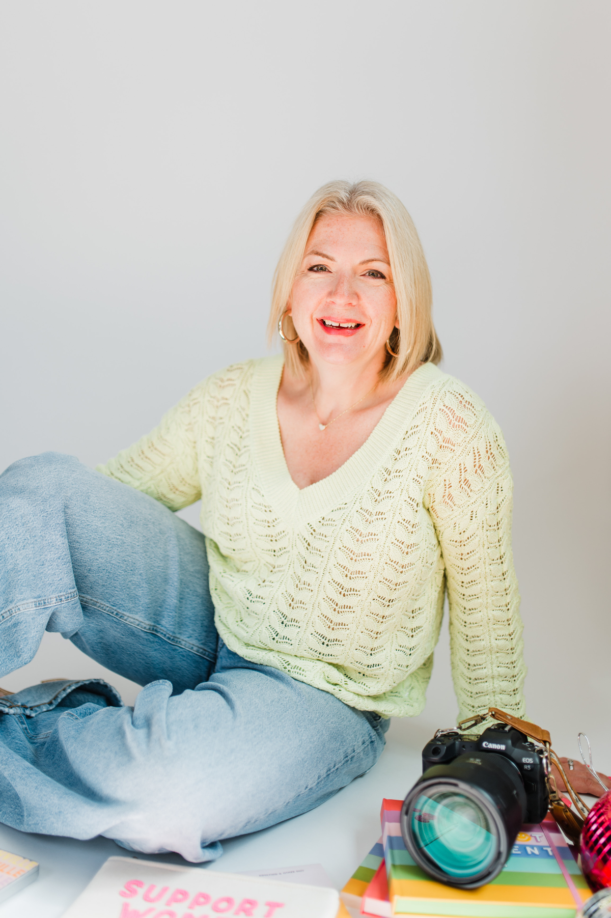 Smiling photography mentor seated on a bright studio floor in a pale green knit sweater and jeans, with a Canon camera resting on colorful books beside her. Clean white backdrop and airy light create a friendly, approachable feel for photography tips for beginners.
