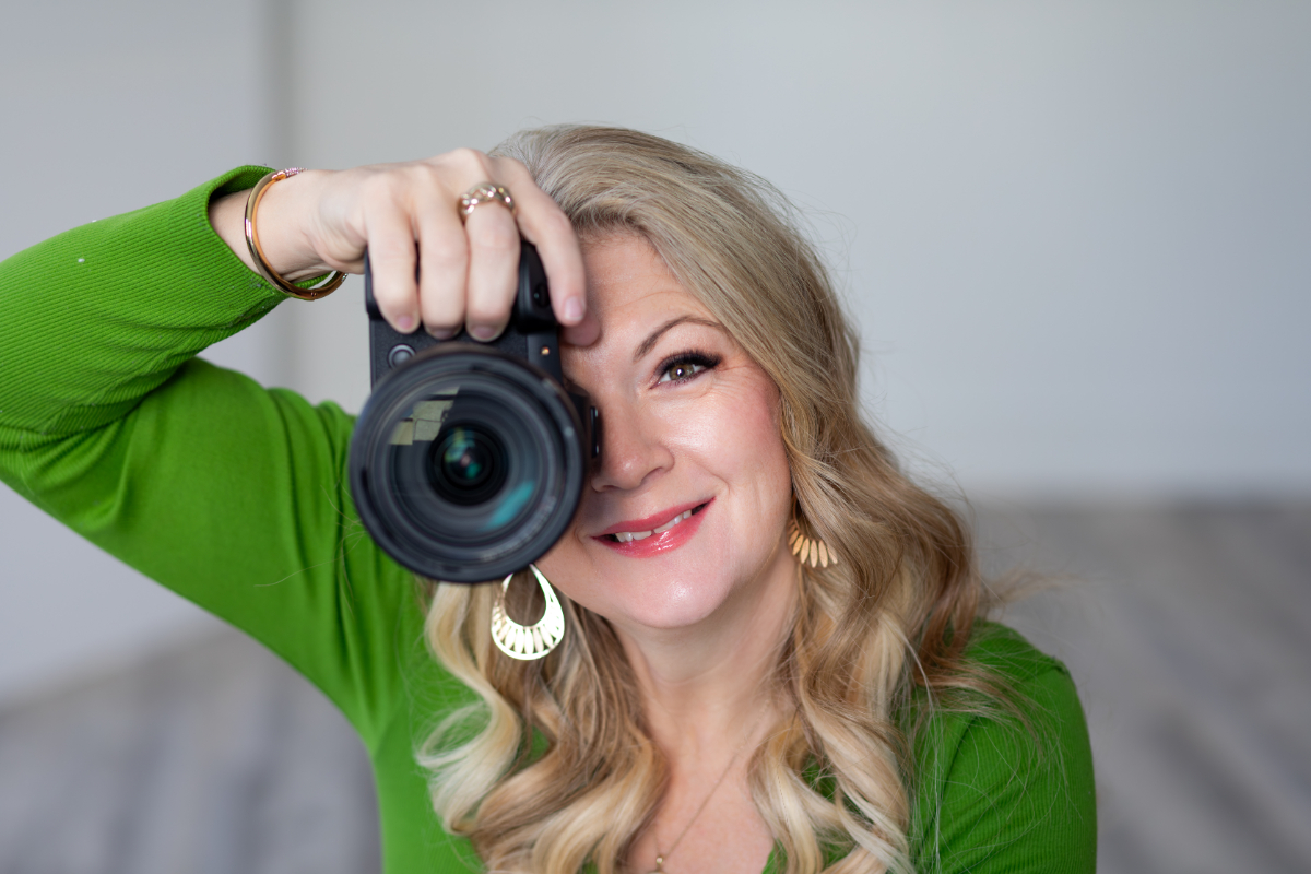 photography-tips-for-beginners Close-up portrait of a blonde photographer in a vivid green top holding a camera to her eye, smiling as she frames a shot. Soft, neutral background and shallow depth of field highlight her face and lens—ideal imagery for photography tips for beginners.