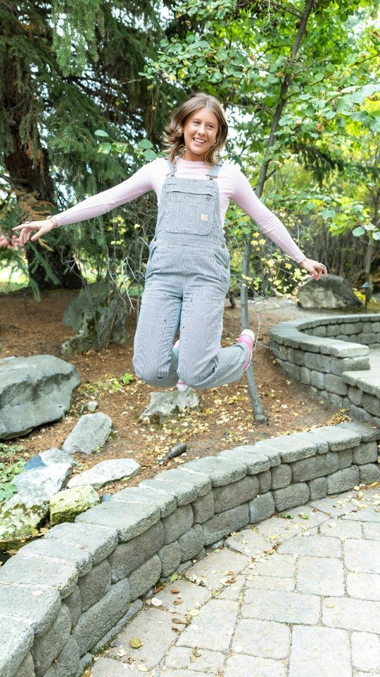 Smiling girl in striped overalls and pink shirt jumps joyfully in a garden setting with stone pathways and trees, captured by a CRM for photographers client session.