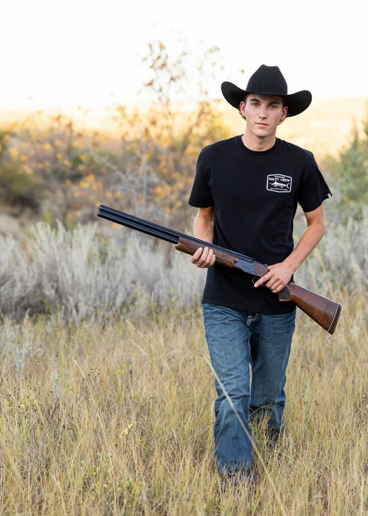 Senior boy in a black cowboy hat and t-shirt walking through tall grass at golden hour, holding a rifle during a CRM for photographers senior session.