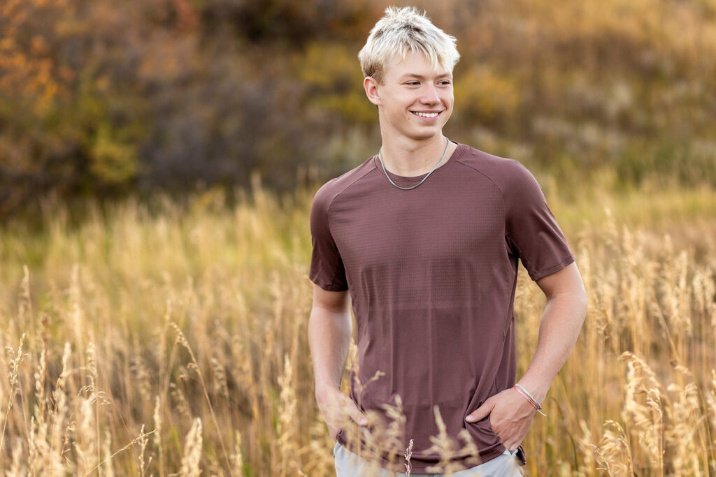 Smiling teenage boy with blonde hair stands in a golden field of tall grass, wearing a maroon t-shirt with his hands in his pockets.