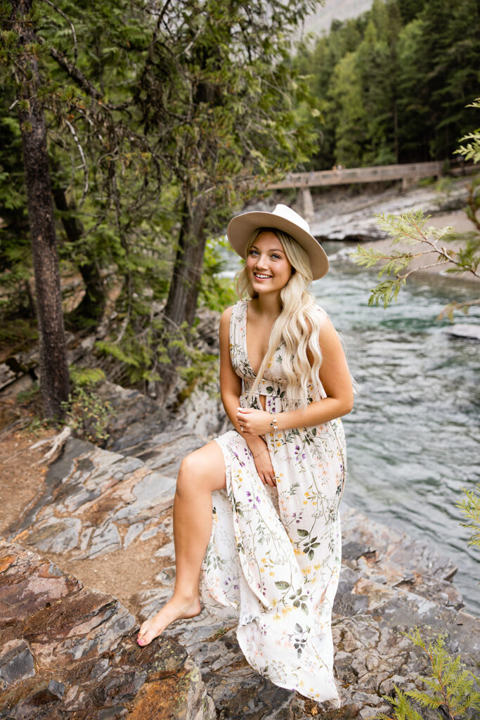 A senior girl in a floral dress smiles next to the water during a GLacier National Park photography session