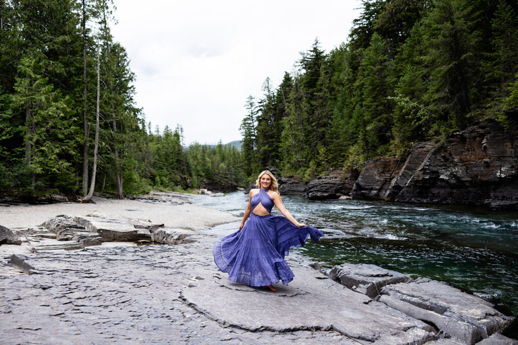A high school senior wearing a purple dress twirls near the water at Glacier National Park for her senior photos in Montana