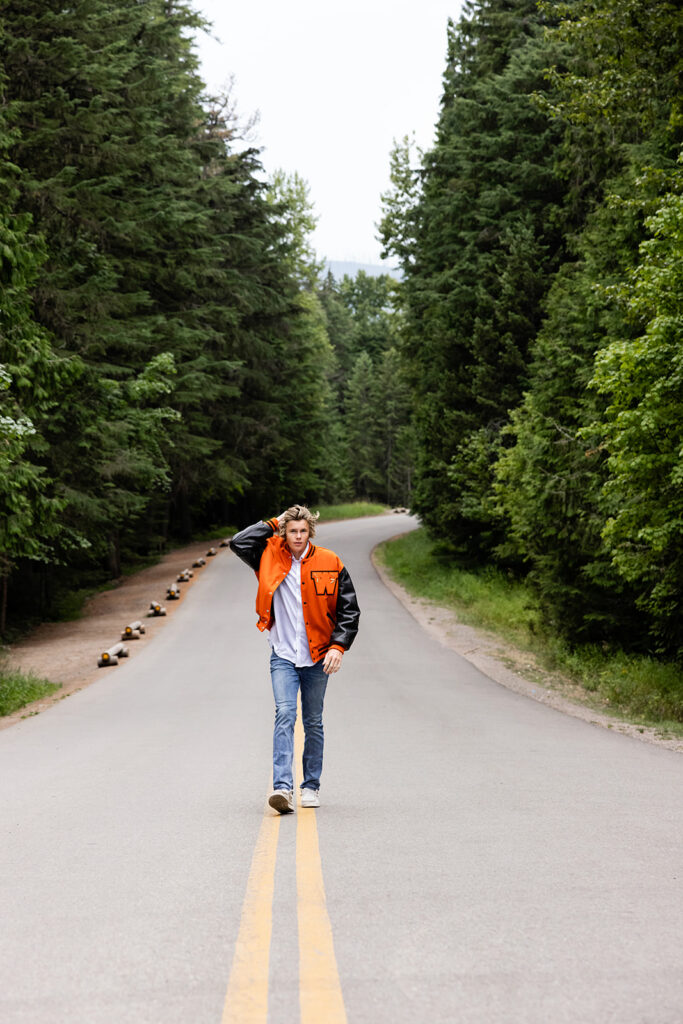 A high school senior boy wearing a letterman's jacket walks down the center of the road in a Glacier National Park Photography session