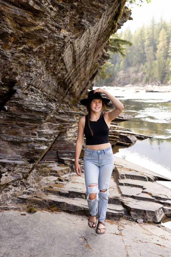 A young girl wearing a black hat and jeans smiles next to the water during her senior photos in Montana at Glacier National Park