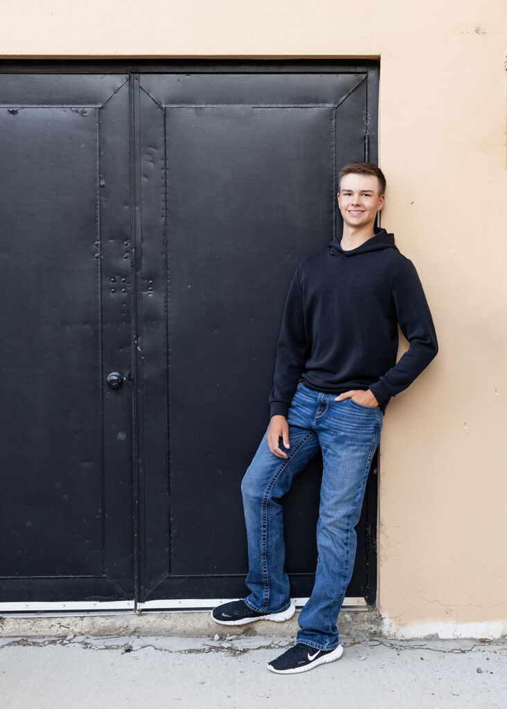 An image from a class of 2026 senior session with North Dakota senior photos depicts a young man in a black sweater & jeans leaning against a wall