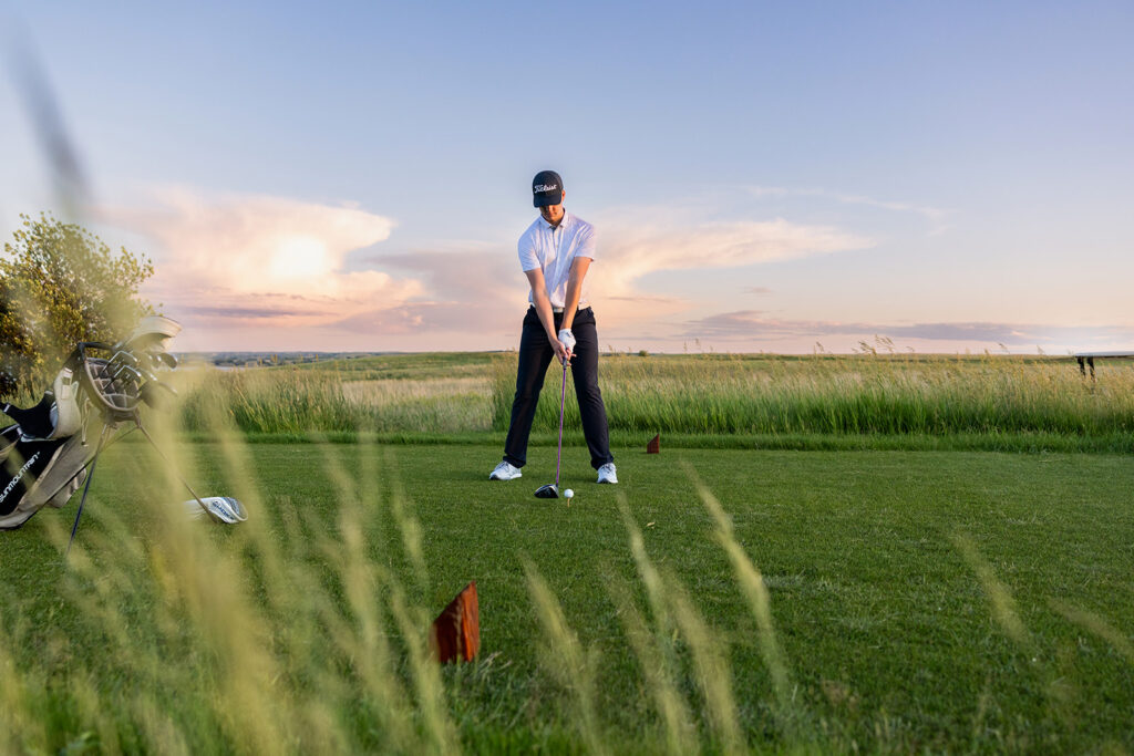 A high school senior boy poses at sunset with his golf clubs during North Dakota senior photos