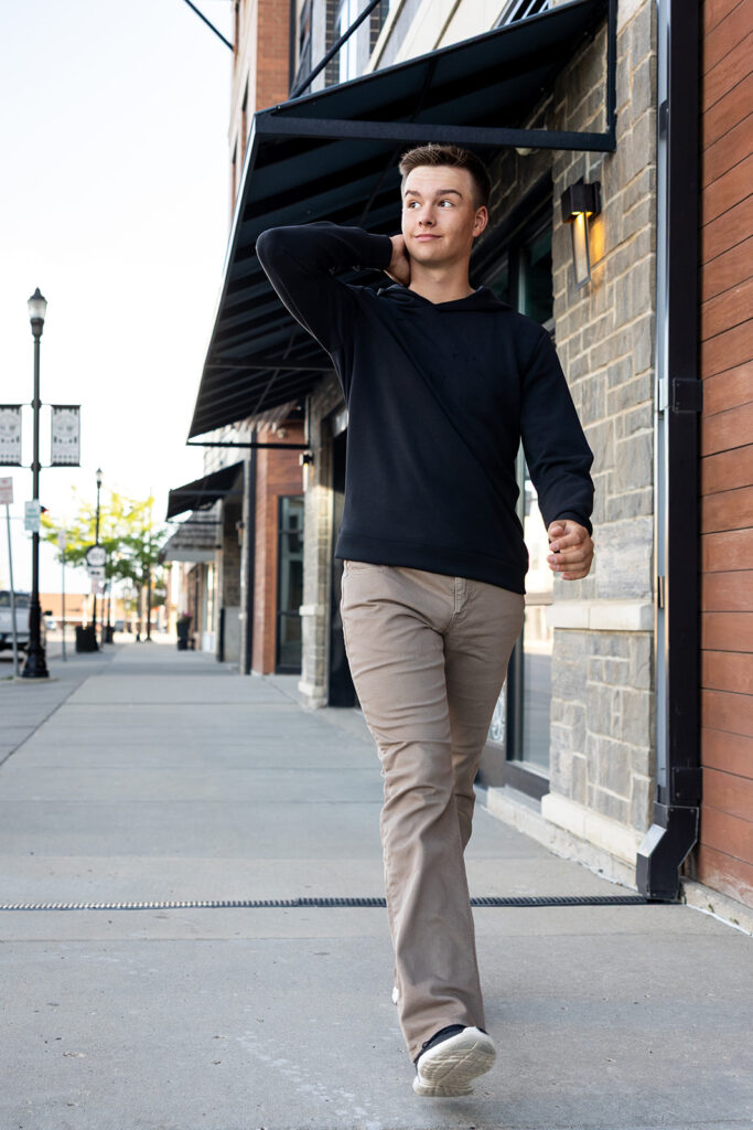 A young man wearing a black sweater and khakis walks down the street of Williston during North Dakota senior photos 