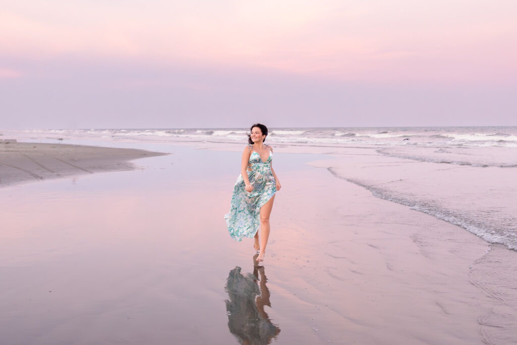 Woman in flowing dress walking barefoot along the shoreline at sunset with soft pastel skies.