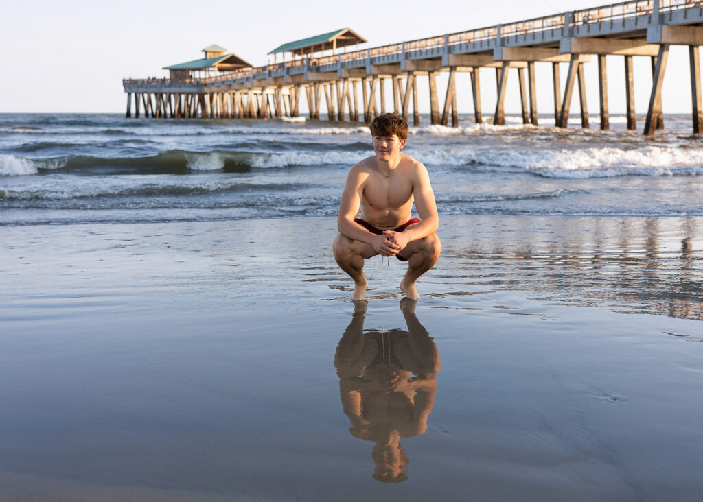 Teen boy crouching on the beach at sunset with pier and reflection in wet sand behind him.