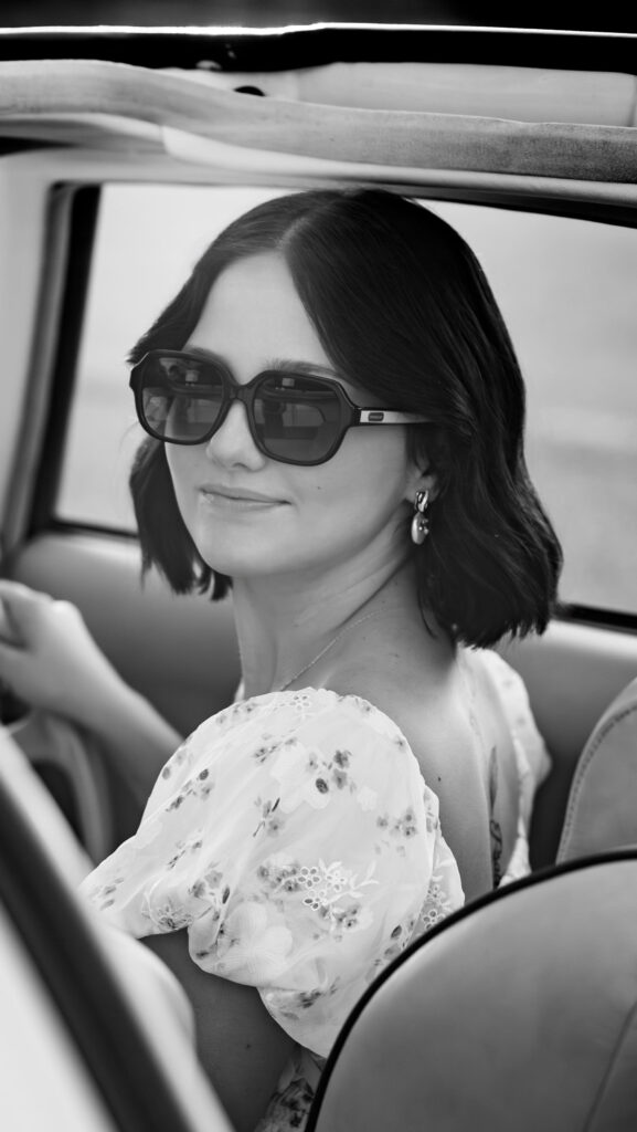 Young woman in sunglasses and a floral dress sitting in a car, looking over her shoulder with a soft smile in a black and white portrait.
