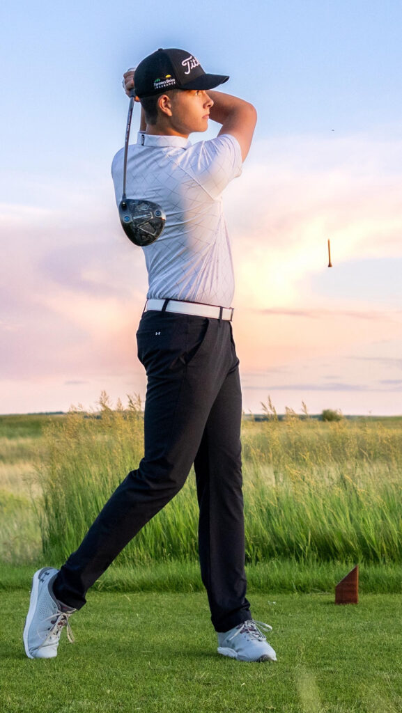 Teen golfer finishing his swing on a green fairway at sunset, with a golf tee flying through the air in the background.