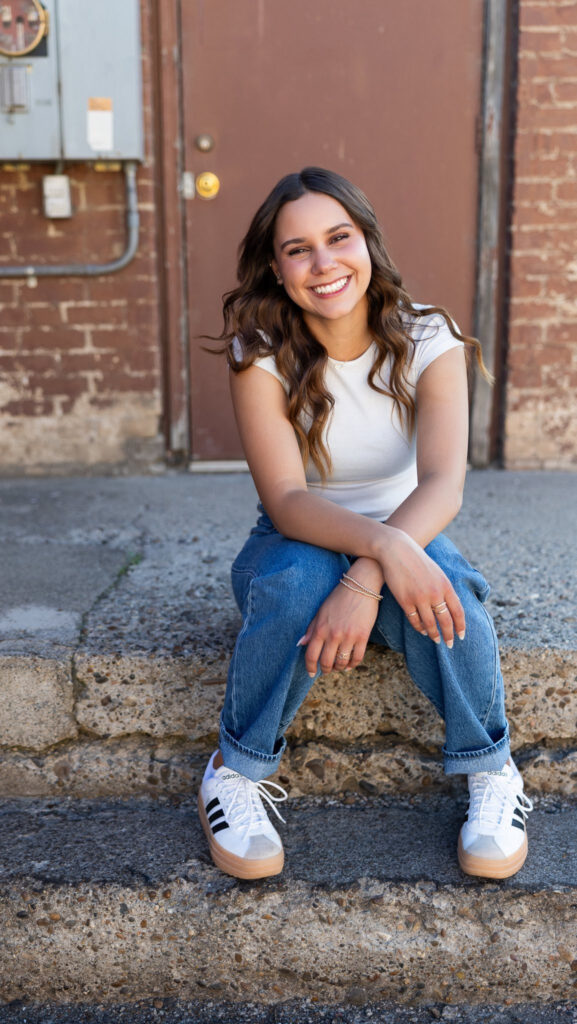 Smiling young woman sitting on concrete steps in a casual outfit with jeans and sneakers, posing in front of a brick building.
