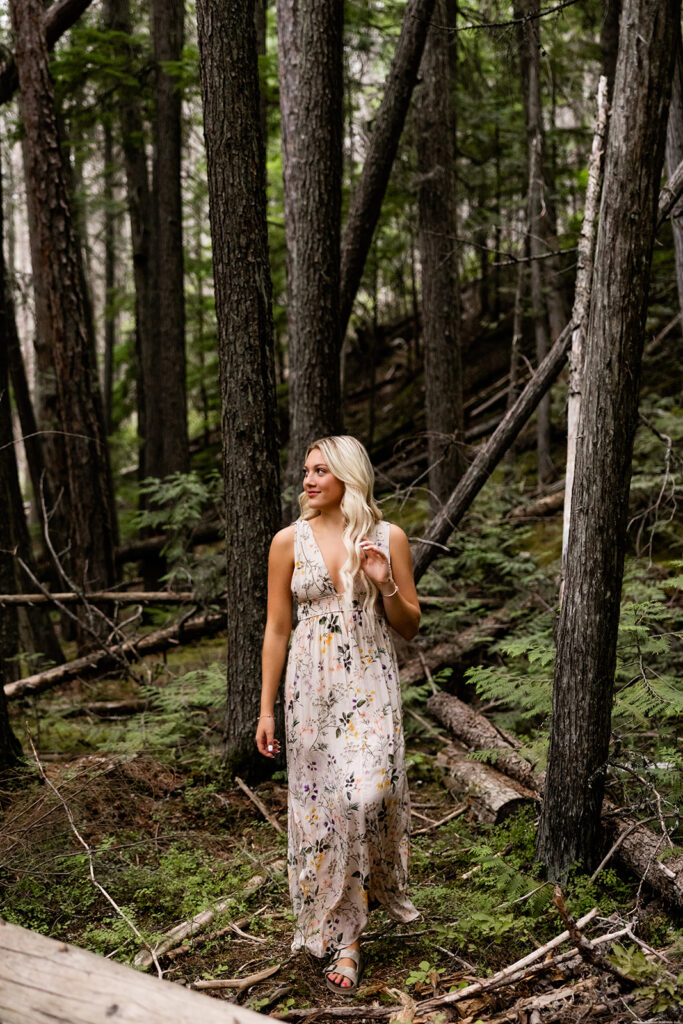 A high school senior in a floor length floral dress walks through the moss covered forest at Glacier National Park during her travel photoshoot with Kellie Rochelle Photography