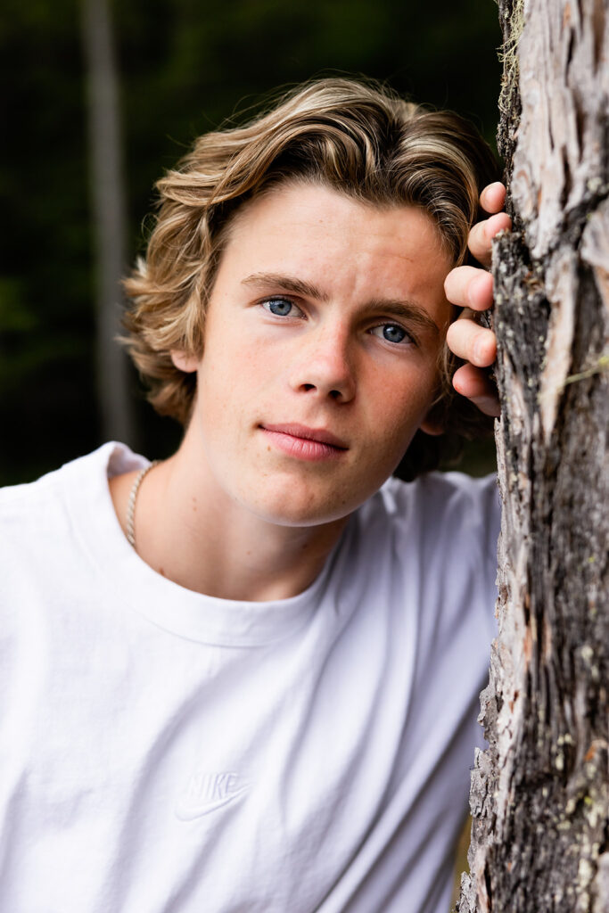 A senior boy wearing a white t lenas against a tree in Glacier National Park in a close-up taken during his senior destination photoshoot