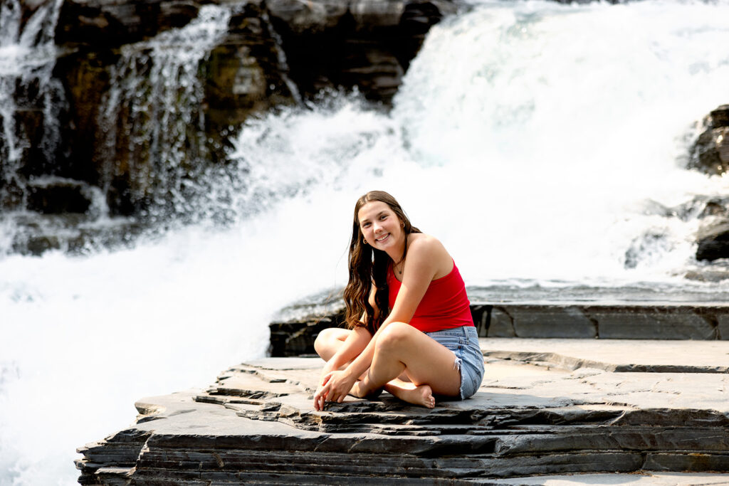 A high school senior girl in jean shorts and a red top smiles from a rock near the river in Glacier National Park during her destination photoshoot with ND senior photographer, Kellie Rochelle Photography