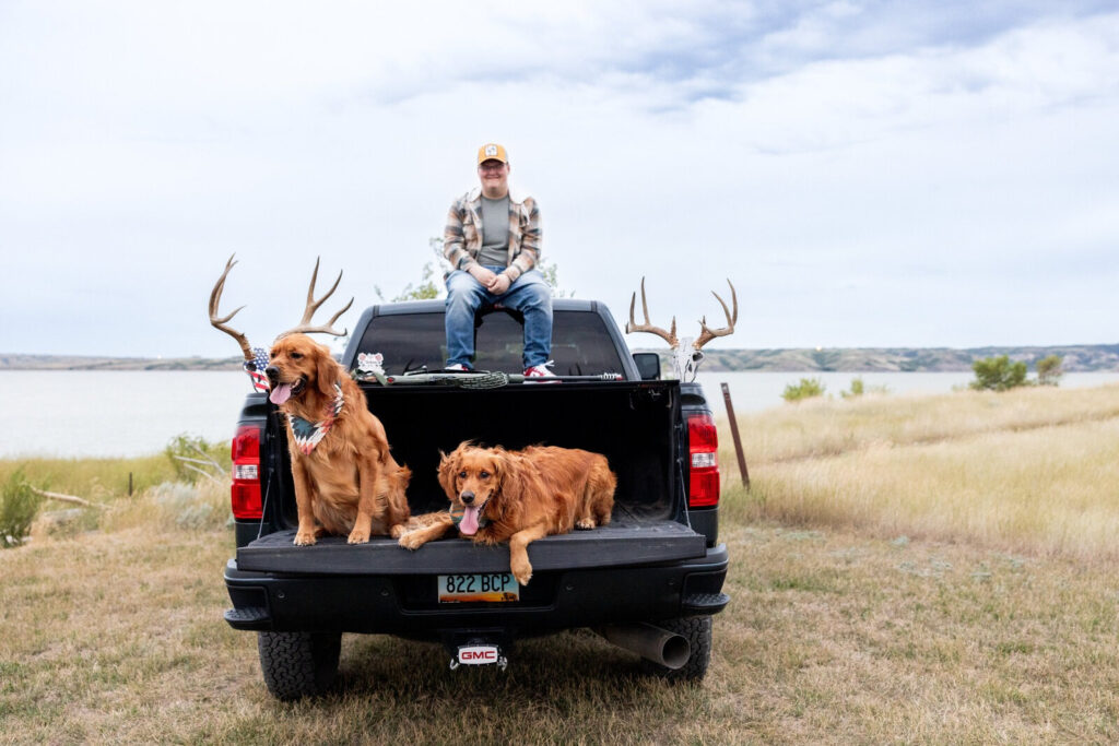 Person sitting on a truck with two golden retrievers and antlers, parked near a lake in open grassland.