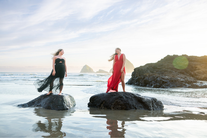Two women in flowing dresses standing on rocks at the beach during sunset, with ocean waves.