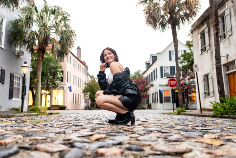Young woman in heels squatting on cobblestone street, surrounded by charming historic buildings and palm trees.