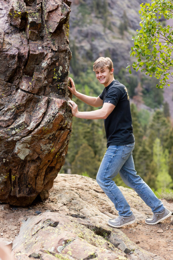 Smiling teenage boy climbing a large rock formation surrounded by forest and mountain scenery.
