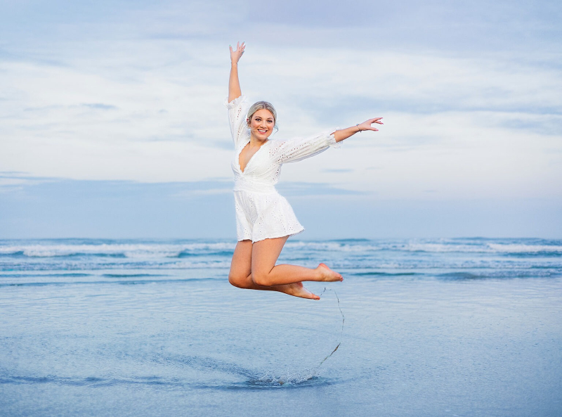 Senior girl jumping joyfully at the beach during off-season travel sessions with ocean backdrop