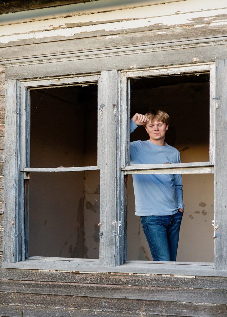 Teen boy leaning in broken window frame during a session at an abandoned rustic location.